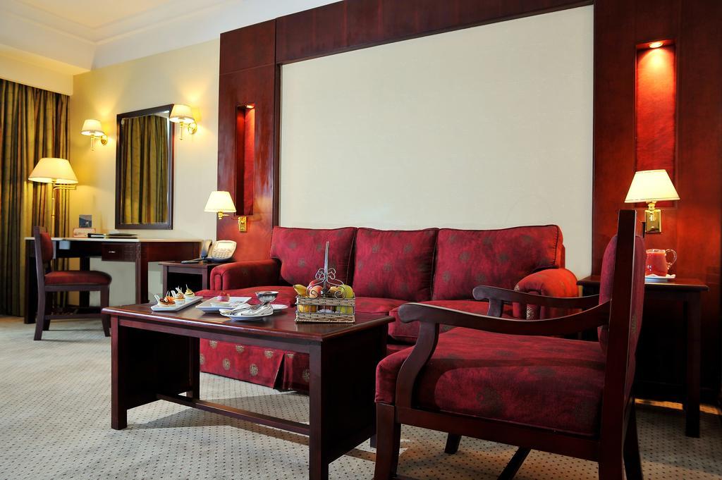 Safir Hotel Cairo Giza Ruang foto The photo shows a well-decorated hotel room or sitting area. In the center, there is a red couch with matching armchairs around it. A wooden coffee table is placed in front of the couch, and it holds some decorative items and snacks. The walls are ad