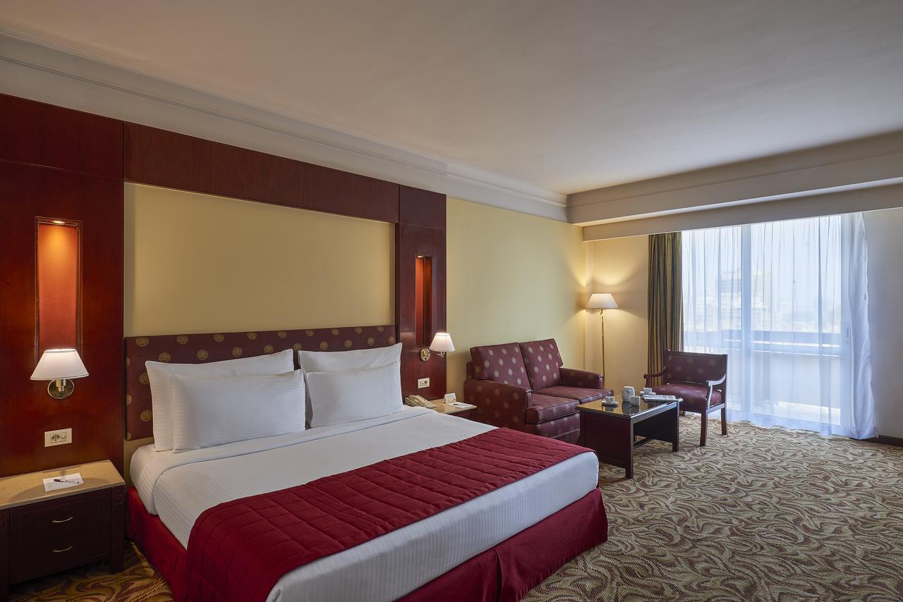 Safir Hotel Cairo Giza Bagian luar foto The image showcases a hotel room featuring a large bed with white linens and a red bedspread. Behind the bed, there is a decorative headboard. The room includes two armchairs and a small coffee table, creating a seating area. Natural light enters thr