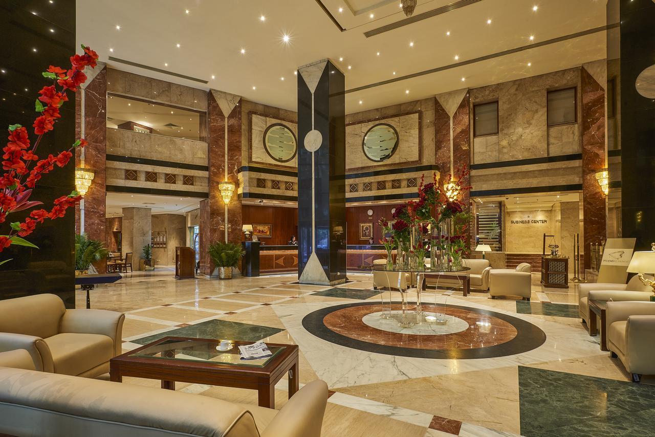 Safir Hotel Cairo Giza Bagian luar foto The photo shows a luxurious hotel lobby. The space features elegant marble flooring with a circular design at the center. There are comfortable seating arrangements with light-colored sofas and wooden coffee tables. Lush green plants and vibrant flor