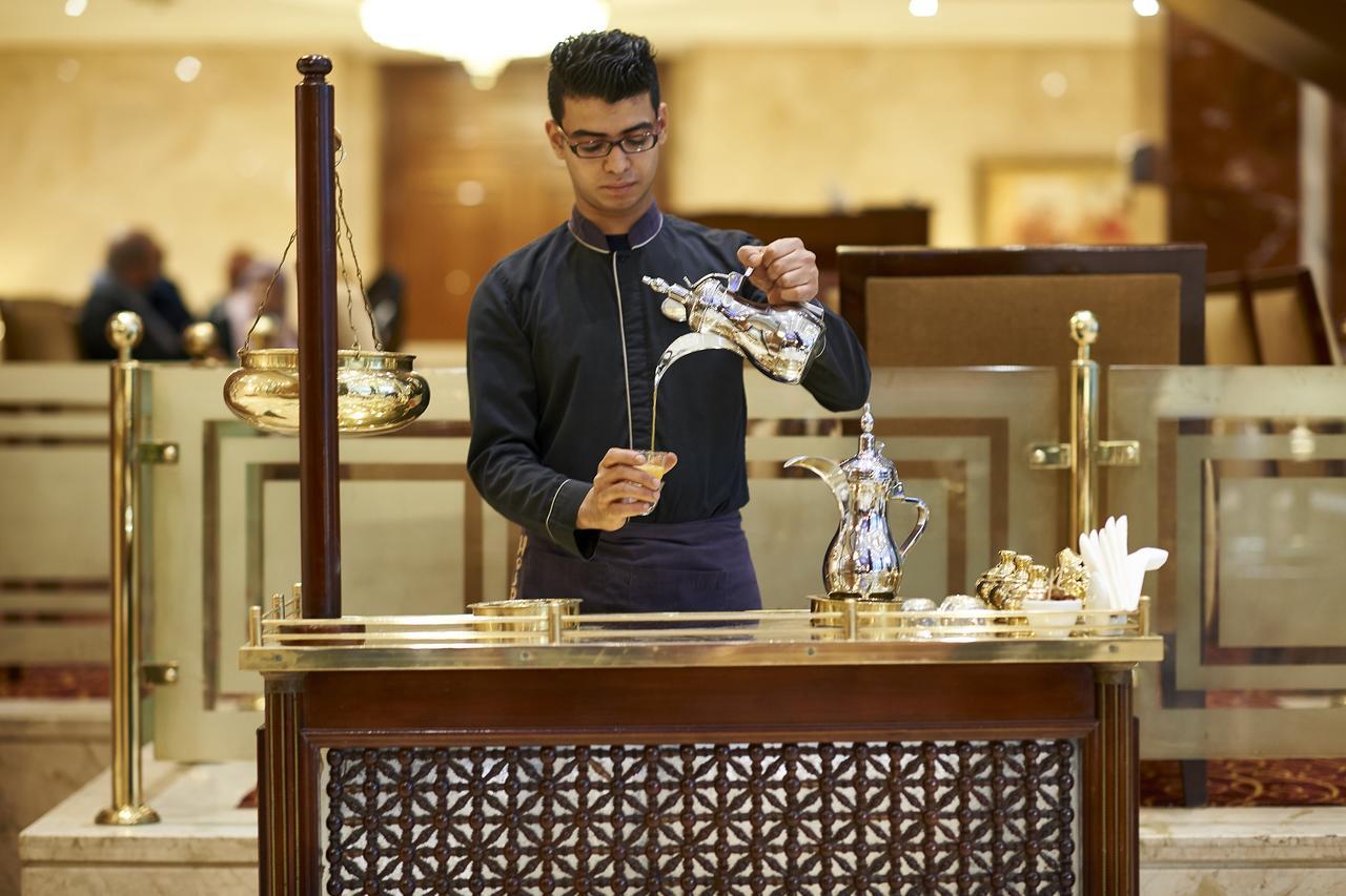 Safir Hotel Cairo Giza Bagian luar foto The photo depicts a person at a service counter, pouring tea from a traditional teapot. The individual appears to be dressed in a formal, dark outfit, and is focused on the task of pouring. The counter features ornate decorations and various tea-serv