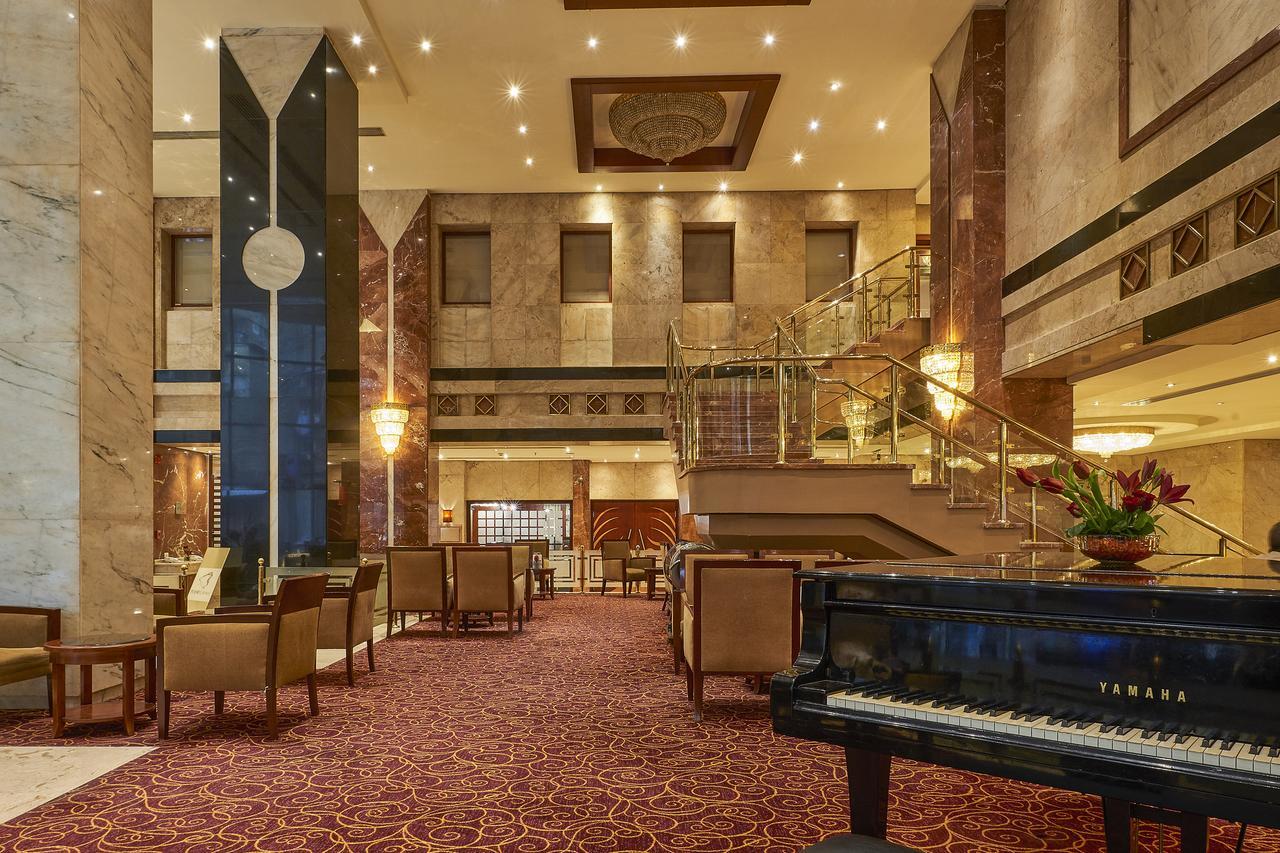 Safir Hotel Cairo Giza Bagian luar foto The image shows an elegant hotel lobby. The space features marble walls and a plush carpet, giving it a luxurious feel. There are seating areas with comfortable chairs arranged around tables. A grand staircase leads to an upper level, and large windo