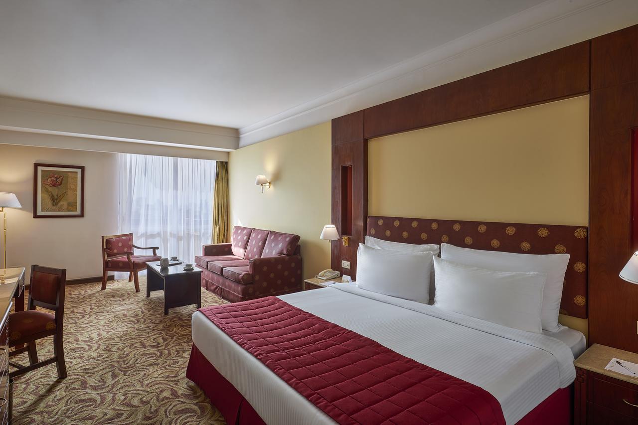 Safir Hotel Cairo Giza Bagian luar foto The photo shows a hotel room featuring a spacious layout. In the center, there is a large bed with white bedding and a red decorative coverlet. To one side, there is a seating area that includes a sofa and two armchairs, all set around a small coffee