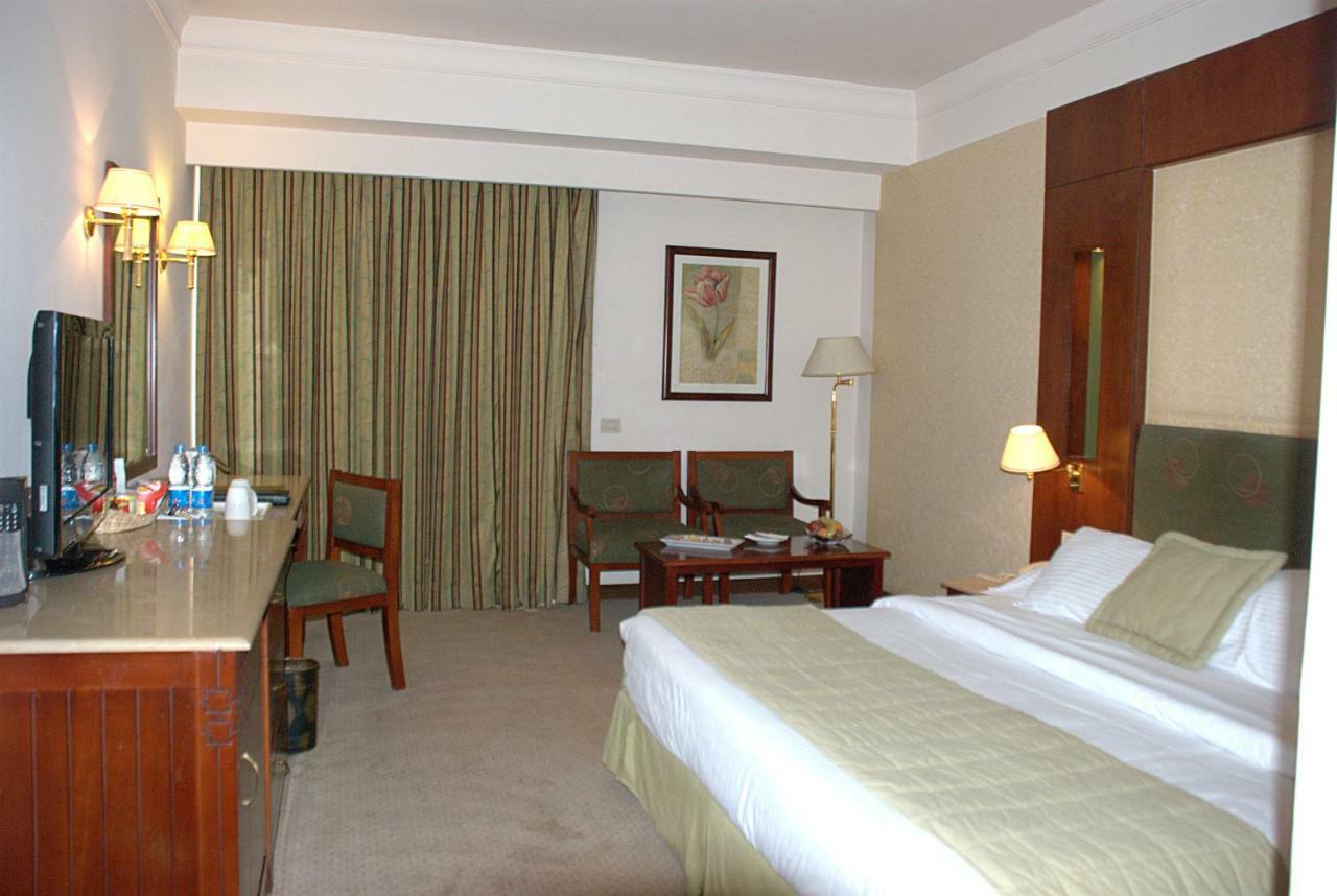 Safir Hotel Cairo Giza Ruang foto The photo shows a hotel room. It features a bed with white linens and a green throw. On one side of the bed, there's a small bedside lamp. Opposite the bed is a desk with a mirror above it, alongside a couple of chairs and a small table. The room has