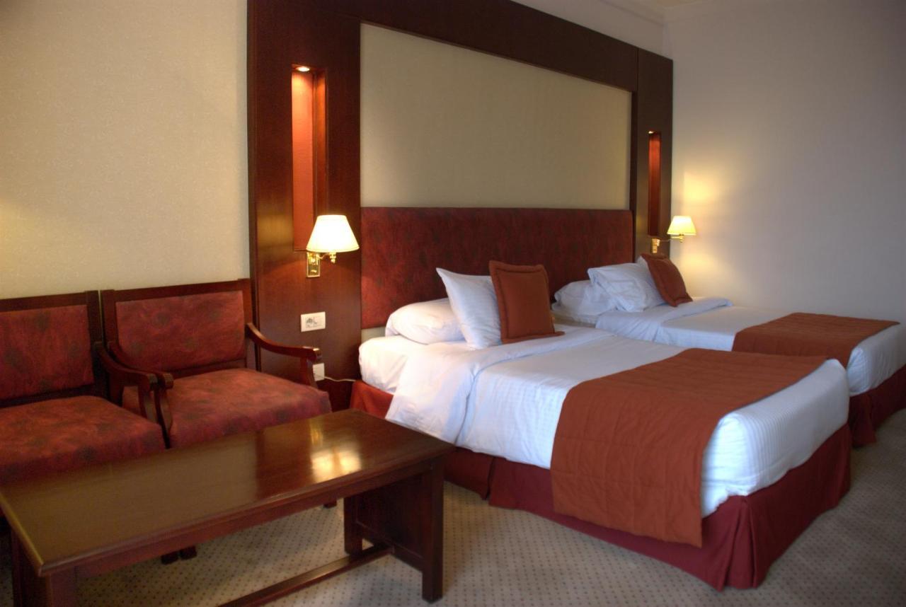 Safir Hotel Cairo Giza Bagian luar foto The photo shows a hotel room featuring two twin beds with white linens and orange bedspreads. There is a headboard behind the beds, along with wall-mounted lamps on either side. In the corner, there are two upholstered chairs and a wooden coffee tabl